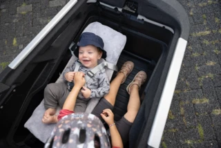 Ein Baby liegt in der Babyschale der Transportbox. Daneben sitzt ein Mädchen im Kindersitz.