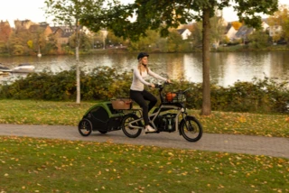 Frau fährt mit beladenem Ca Go CS an einem Fluss entlang. Am Rad ist ein Anhänger befestigt, in dem ein Hund sitzt.