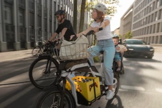 Frau fährt mit Ca Go CS durch die Stadt. Auf dem Gepäckträger ist ein Kindersitz montiert, in dem ein kleines Kind sitzt. Neben der Frau fährt ein Mann mit einem E-Bike.