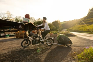Frau fährt mit beladenem Ca Go CS auf einen Hof. Am Rad ist ein Anhänger befestigt, in dem ein Hund sitzt.