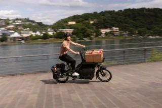 Frau fährt mit CS Vario an einem Fluss. Das Extended Front Rack ist mit Kartons beladen.