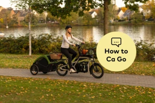 Frau fährt mit beladenem Ca Go CS an einem Fluss entlang. Am Rad ist ein Anhänger befestigt, in dem ein Hund sitzt.